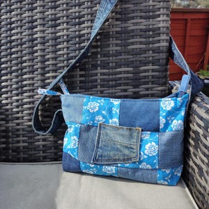May include: A denim patchwork shoulder bag featuring a blue floral pattern and a denim pocket. The bag has a long denim strap and is made from various shades of blue denim. The bag is on a grey wicker chair.