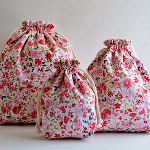 May include: Three drawstring bags made of white fabric with a pink and red floral pattern. The bags are different sizes and are arranged in a row from largest to smallest.