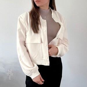May include: A white cropped bomber jacket with a zipper closure and a button-up front. The jacket has two large pockets on the front and a ribbed hem and cuffs.