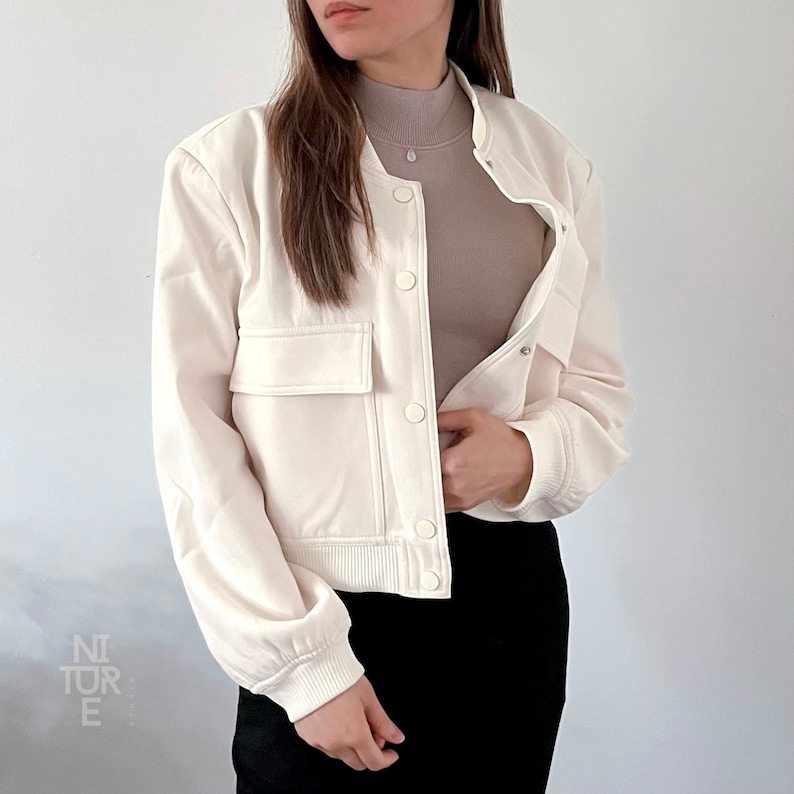 May include: A white cropped bomber jacket with a zipper closure and a button-up front. The jacket has two large pockets on the front and a ribbed hem and cuffs.