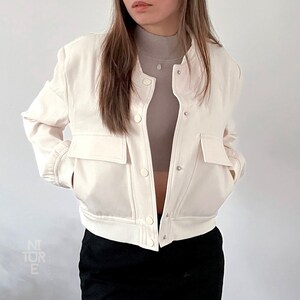 May include: A white cropped bomber jacket with a zipper closure and two front pockets. The jacket has a ribbed hem and cuffs.
