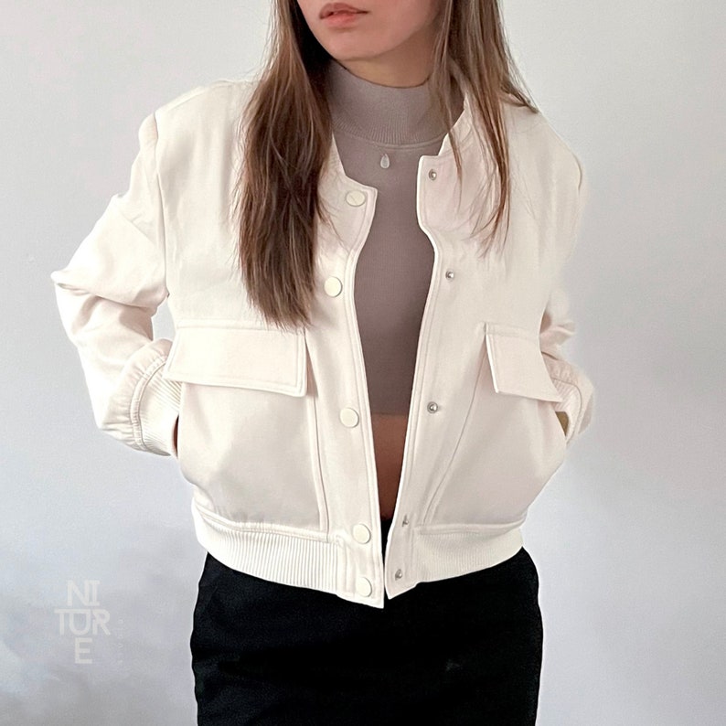 May include: A white cropped bomber jacket with a zipper closure and two front pockets. The jacket has a ribbed hem and cuffs.
