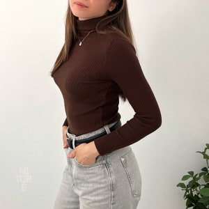 Peut inclure: Un pull à col roulé côtelé marron à manches longues. Le pull est associé à un jean gris clair et une ceinture noire.