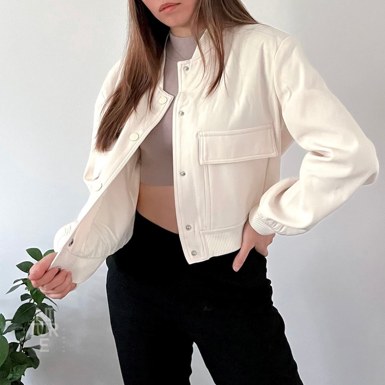 May include: A white cropped bomber jacket with a zipper closure and a large pocket on the left side. The jacket has a ribbed hem and cuffs.