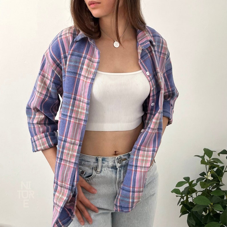 Może przedstawiać: Koszula flanelowa w niebiesko-r&oacute;żową kratę z białym tank topem pod spodem. Koszula jest rozpięta, a tank top jest widoczny. Osoba nosi niebieskie dżinsy.