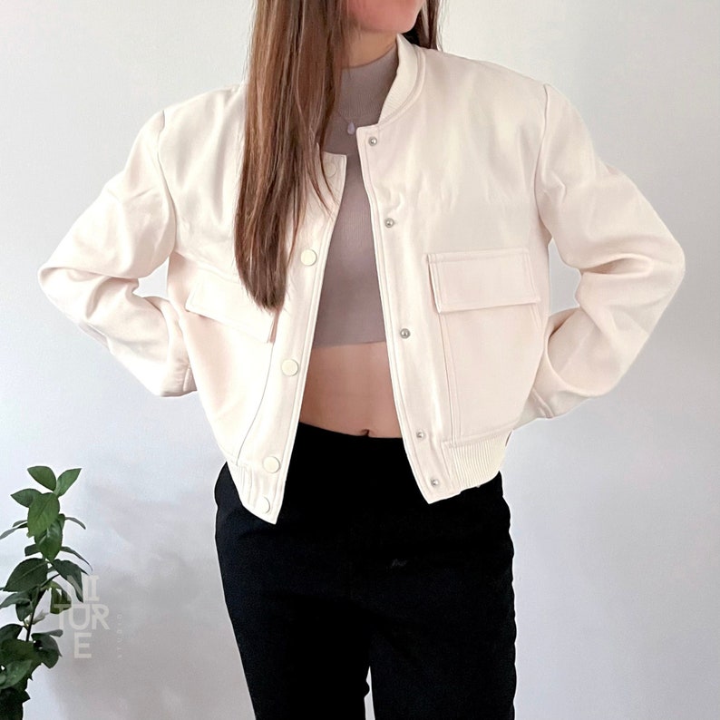 May include: A white cropped bomber jacket with a zipper closure and a large pocket on the left side. The jacket has a ribbed hem and cuffs.