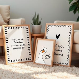 May include: Three framed art prints with light wood frames. One print has the text "Ici, on se lave les mains... et la conscience". Another has a black heart and text. The third features a cartoon goose with the text "Joli cul".