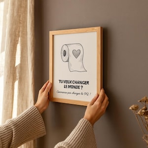 K&ouml;nnte beinhalten: Ein gerahmter Druck mit einem hellen Holzrahmen. Der Druck zeigt eine Schwarz-Wei&szlig;-Illustration einer Toilettenpapierrolle mit einem Herz. Der Text lautet: &bdquo;TU VEUX CHANGER LE MONDE? Commence par changer le PQ!"