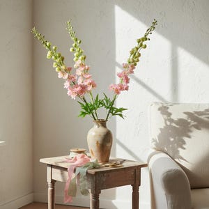 May include: A rustic wooden side table holds a beige ceramic vase filled with pink and green flowers. Ribbons in pastel colors and a pair of scissors rest on the table. The scene is set against a white wall with sunlight.