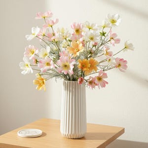 Könnte beinhalten: Ein Strauß Kosmosblumen in einer weißen, geriffelten Vase. Die Blumen sind in Rosa, Gelb und Weiß gehalten, mit grünen Stielen. Die Vase steht auf einem hellen Holztisch mit einem Untersetzer.