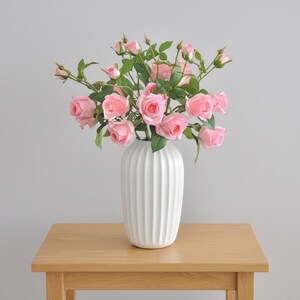 Può includere: Un vaso di ceramica bianco e scanalato pieno di rose rosa e foglie verdi è posto su un tavolo di legno marrone chiaro. Le rose sono in piena fioritura, con alcuni boccioli visibili. Lo sfondo è un muro grigio chiaro.