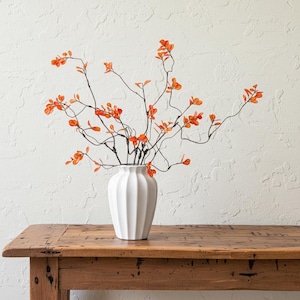 Puede incluir: Un jarrón de cerámica blanco acanalado que contiene ramas con hojas naranjas. El jarrón está sobre una mesa de madera rústica contra una pared blanca texturizada. Las ramas tienen tallos finos y oscuros y hojas de color naranja vibrante.