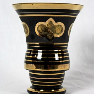 May include: A black glass vase with gold accents. The vase has a wide mouth and a narrow base. It is decorated with gold stripes and a gold fleur-de-lis design.