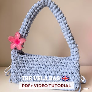 May include: A light blue crocheted handbag with a rectangular body and a curved handle. A pink flower is attached to the handle. The text "THE VELA BAG PDF+ VIDEO TUTORIAL" is displayed on the bag.