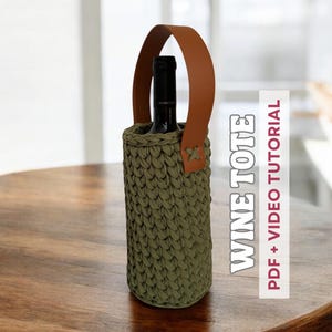 May include: A green crocheted wine tote bag with a brown leather handle. The tote bag is sitting on a wooden table.  The text "WINE TOTE" is printed on the side of the bag. The text "PDF + VIDEO TUTORIAL" is printed on the right side of the image.
