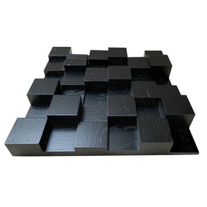 Puede incluir: Un panel de pared de madera negra con un diseño geométrico. El panel está compuesto por numerosos bloques cuadrados de diferentes alturas, creando un efecto texturizado y tridimensional. La veta de la madera es visible.