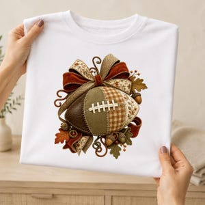Könnte beinhalten: Ein weißes T-Shirt mit einer zentralen Grafik eines Footballs, der mit verschiedenen strukturierten Mustern in Braun-, Olivgrün- und Cremetönen gestaltet ist. Der Football ist mit einer großen dekorativen Schleife, Herbstblättern und kleinen Eicheln verziert.