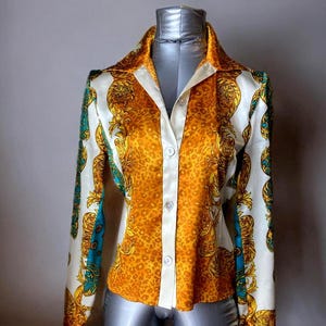 May include: A button-down shirt with a vibrant design. The shirt features a white collar and button placket, with a bold pattern of gold and orange animal print and ornate gold detailing. The sleeves have a white background with gold and teal accents.