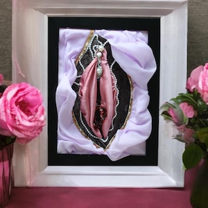 Può includere: Opera d'arte incorniciata raffigurante genitali femminili in tessuto. Il pezzo è incorniciato in bianco, con un bordo interno nero. L'opera d'arte utilizza tessuti rosa, neri e bianchi, con accenti di perle e perline. Rose rosa sono visibili a sinistra e a destra.