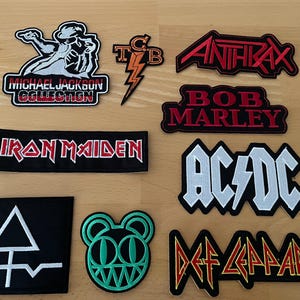 May include: A collection of embroidered fabric patches featuring various music artists and logos, including Michael Jackson, TCB, Anthrax, Bob Marley, Iron Maiden, AC/DC, a geometric symbol, a green bear face, and the Def Leppard band logo.