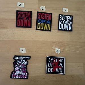 Peut inclure: Cinq écussons avec le texte "SYSTEM OF A DOWN" dans différents designs. Les écussons présentent du texte rouge, bleu et blanc sur des fonds noirs. Un écusson a un motif de crâne. Les écussons sont carrés et rectangulaires.