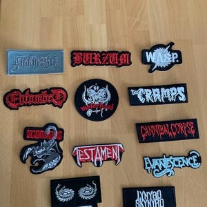Könnte beinhalten: Eine Sammlung von aufgestickten Aufnähern mit verschiedenen Bandlogos und -namen. Die Aufnäher sind in verschiedenen Formen und Größen gehalten, mit einer Mischung aus Farben wie Schwarz, Rot, Weiß und Blau. Die Aufnäher enthalten Bandnamen wie "Burzum", "WASP" und "Cannibal Corpse".