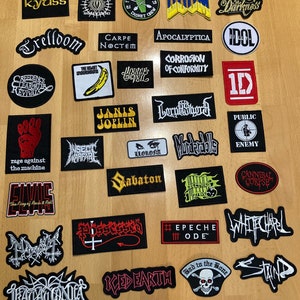 Può includere: Una collezione di toppe ricamate con loghi di band e testo. Le toppe includono loghi per Kyuss, Rage Against the Machine, Janis Joplin e altri. Le toppe sono di varie forme e colori, con testo in bianco, giallo e rosso.