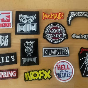 Peut inclure: Une collection d'écussons en tissu brodé de formes rectangulaires, circulaires et variées, présentant des logos de groupes et du texte, notamment Limp Bizkit, Motionless In White, Mac Miller, Killswitch Engage, Incubus, Amon Amarth, Good Charlotte, Pixies, Beartooth, Kilmister, The Offspring, NOFX, To Hell with the Beatles et Metallica.
