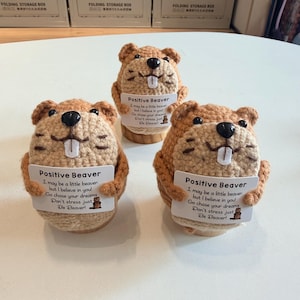 Könnte beinhalten: Drei gehäkelte Biberfiguren in Braun- und Beigetönen, die jeweils ein Schild mit der Aufschrift "Positive Beaver" halten. Die Schilder enthalten eine inspirierende Botschaft und eine kleine Biberillustration. Die Biber werden auf Holzständern präsentiert.