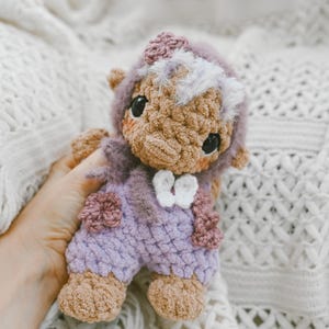 May include: A handmade crochet doll in shades of brown, lavender, and white. The doll has a fuzzy head covering, large black eyes, and is wearing a lavender outfit with floral accents. The doll is approximately 6 inches tall.