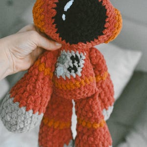 May include: Handmade crochet doll in shades of red, orange, and gray. The doll has a large black face with white accents, and a small patch on the chest. The arms and legs are short and stubby, and the doll is made of soft yarn.