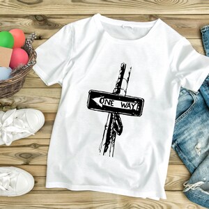 K&ouml;nnte beinhalten: Wei&szlig;es T-Shirt mit einem schwarz-wei&szlig;en "ONE WAY" Stra&szlig;enschild-Motiv. Das Bild zeigt wei&szlig;e Turnschuhe, einen Korb mit bunten Eiern und eine blaue Jeans auf einer Holzoberfl&auml;che.