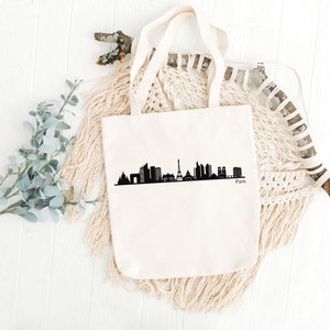 May include: A white canvas tote bag with a black silhouette of the Paris skyline, including the Eiffel Tower, and the word "Paris" printed below.