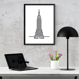 K&ouml;nnte beinhalten: Ein gerahmter Schwarz-Wei&szlig;-Druck des Empire State Building mit dem Text "New York" darunter. Der Druck wird auf einem wei&szlig;en Schreibtisch mit einer schwarzen Schreibtischlampe, einem Laptop und einem kleinen wei&szlig;en Topf mit rosa Blumen angezeigt.