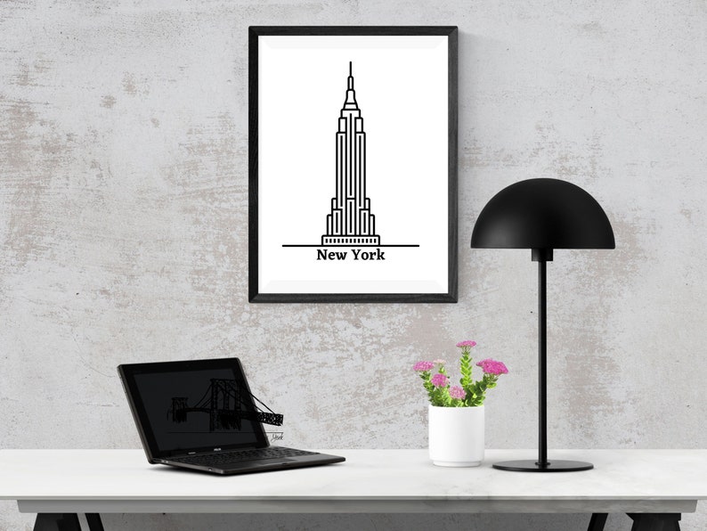 K&ouml;nnte beinhalten: Ein gerahmter Schwarz-Wei&szlig;-Druck des Empire State Building mit dem Text "New York" darunter. Der Druck wird auf einem wei&szlig;en Schreibtisch mit einer schwarzen Schreibtischlampe, einem Laptop und einem kleinen wei&szlig;en Topf mit rosa Blumen angezeigt.