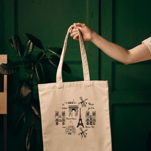 May include: A white canvas tote bag with black illustrations of Parisian landmarks, including the Eiffel Tower, Notre Dame, and the Arc de Triomphe. The text "Je t'aime Paris" is printed on the bag.