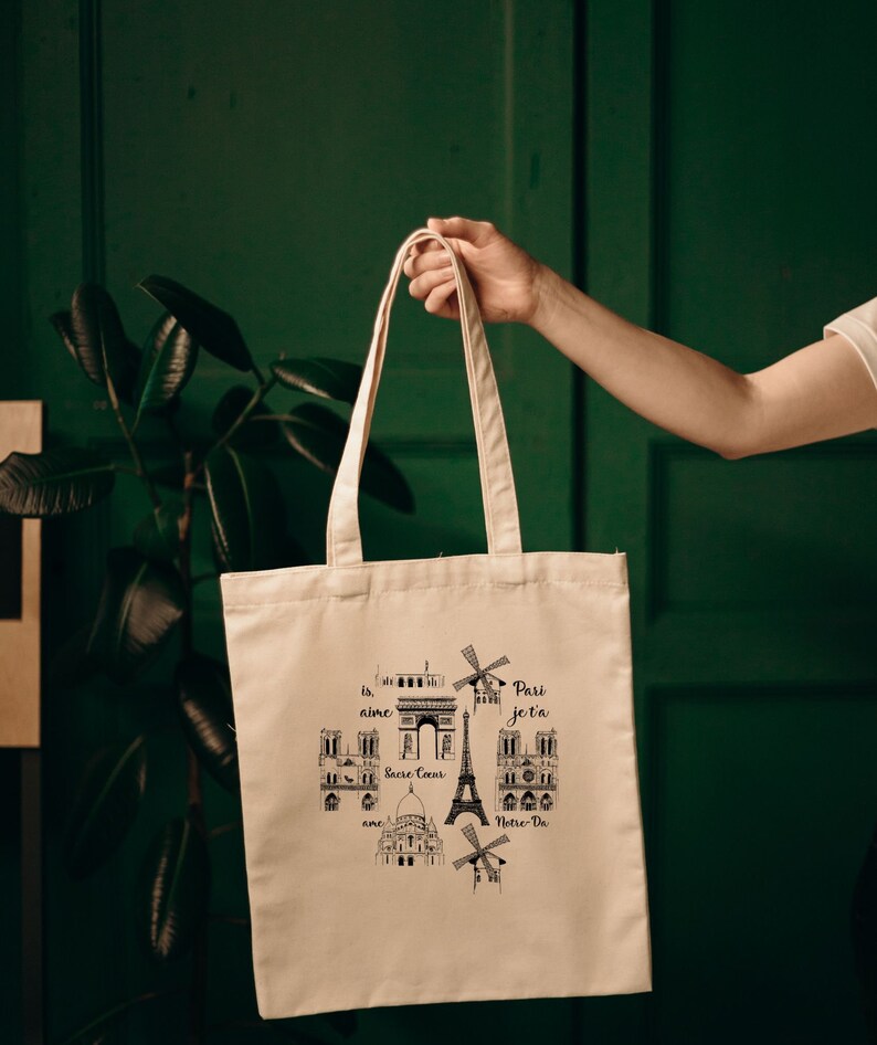 May include: A white canvas tote bag with black illustrations of Parisian landmarks, including the Eiffel Tower, Notre Dame, and the Arc de Triomphe. The text "Je t'aime Paris" is printed on the bag.