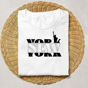 K&ouml;nnte beinhalten: Wei&szlig;es T-Shirt mit schwarzem "NEW YORK"-Schriftzug und der Silhouette der Freiheitsstatue. Das gefaltete Shirt liegt auf einer runden, gewebten Unterlage.