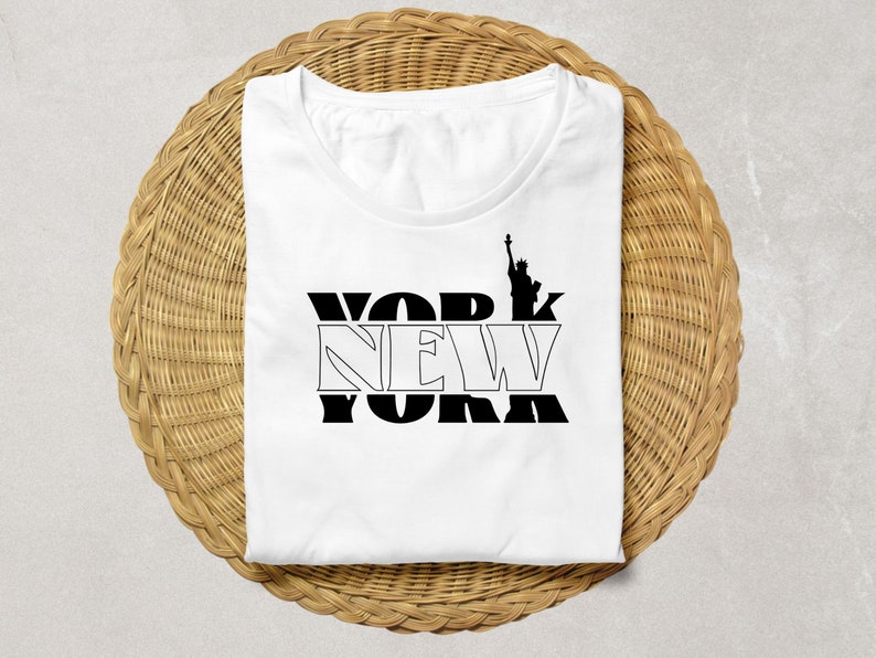 K&ouml;nnte beinhalten: Wei&szlig;es T-Shirt mit schwarzem "NEW YORK"-Schriftzug und der Silhouette der Freiheitsstatue. Das gefaltete Shirt liegt auf einer runden, gewebten Unterlage.
