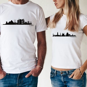 May include: Two white t-shirts with black city skyline silhouettes. The shirt on the left has the text "CAIRO" below the skyline. The shirt on the right has the text "CASABLANCA" below the skyline.