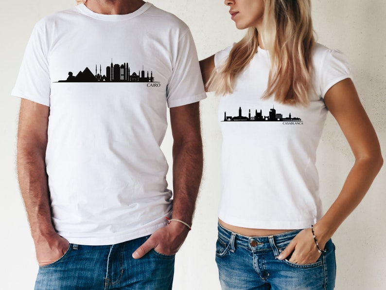 May include: Two white t-shirts with black city skyline silhouettes. The shirt on the left has the text "CAIRO" below the skyline. The shirt on the right has the text "CASABLANCA" below the skyline.