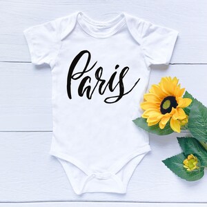 May include: White baby onesie with the word "Paris" written in black cursive script. The onesie is lying on a white wooden surface with a yellow sunflower.