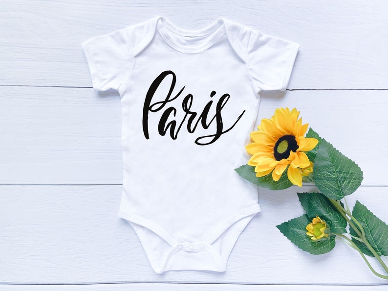 May include: White baby onesie with the word "Paris" written in black cursive script. The onesie is lying on a white wooden surface with a yellow sunflower.