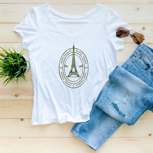 May include: White v-neck t-shirt with a graphic of the Eiffel Tower and the text "Eiffel Tower, Paris, France". The shirt is laid flat on a wooden surface with a pair of blue jeans and sunglasses.