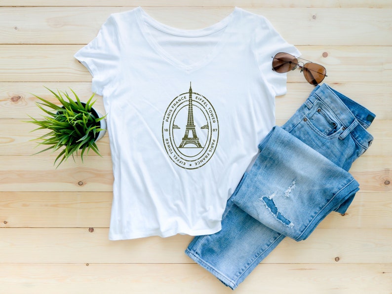 May include: White v-neck t-shirt with a graphic of the Eiffel Tower and the text "Eiffel Tower, Paris, France". The shirt is laid flat on a wooden surface with a pair of blue jeans and sunglasses.