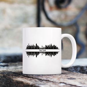 K&ouml;nnte beinhalten: Wei&szlig;e Keramiktasse mit einer schwarzen Silhouette der Skyline von New York City. Das Design zeigt die Wahrzeichen der Stadt und den Namen "New York". Die Tasse hat einen gebogenen Griff.