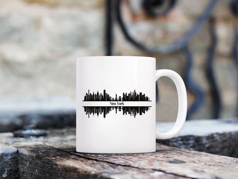 K&ouml;nnte beinhalten: Wei&szlig;e Keramiktasse mit einer schwarzen Silhouette der Skyline von New York City. Das Design zeigt die Wahrzeichen der Stadt und den Namen "New York". Die Tasse hat einen gebogenen Griff.