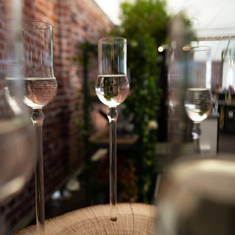 May include: Four clear glass shot glasses with a wooden surface in the background. The glasses are filled with a clear liquid and reflect the surrounding environment.