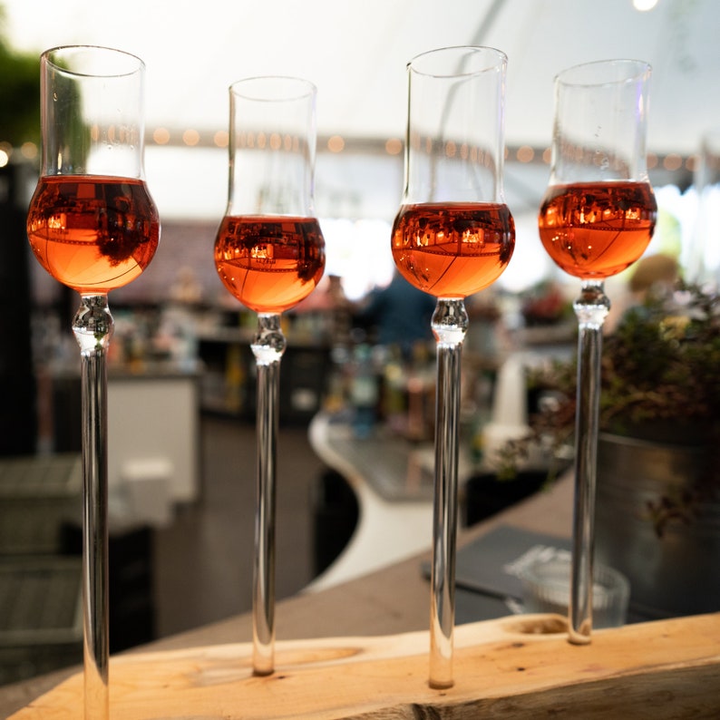 May include: Four clear glass shot glasses filled with a light orange liquid, sitting on a wooden surface. The glasses are arranged in a row, with the stems of the glasses extending down to the surface.