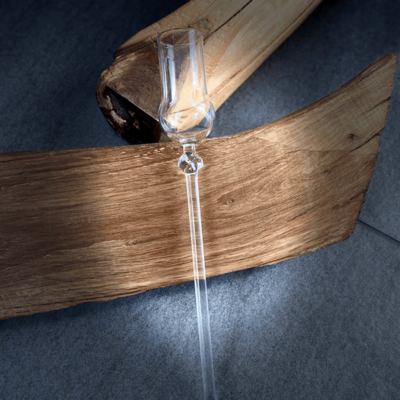May include: A clear glass shot glass with a long, thin stem. The glass is resting on a piece of wood.